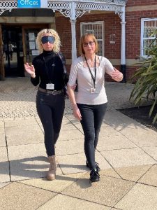 This is a training activity showing two adults, one is wearing sleep shades and is being guided by the sighted adult by holding on to their arm above the elbow. They are walking outside on a bright sunny day. The sighted guide is walking half a step ahead of the adult wearing sleep shades.