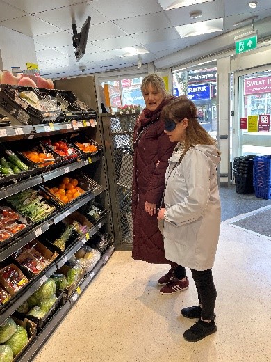 This is a training activity showing two adults, one is wearing sleep shades and is being guided by the sighted adult. They are inside a grocery store, the sighted guide is giving a description of they types of fruits and vegetables on display.